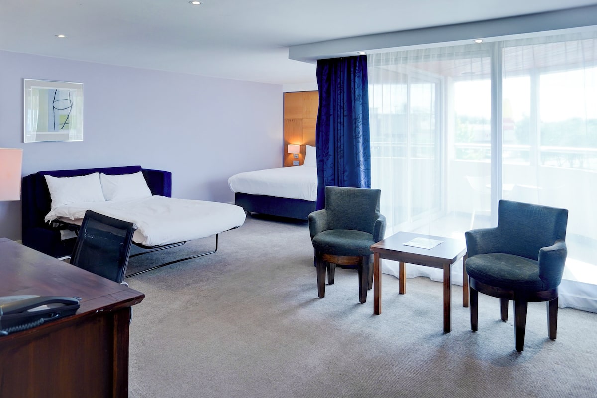 Una habitación de hotel con una cama, dos sillones, un escritorio y un ventanal con cortinas.