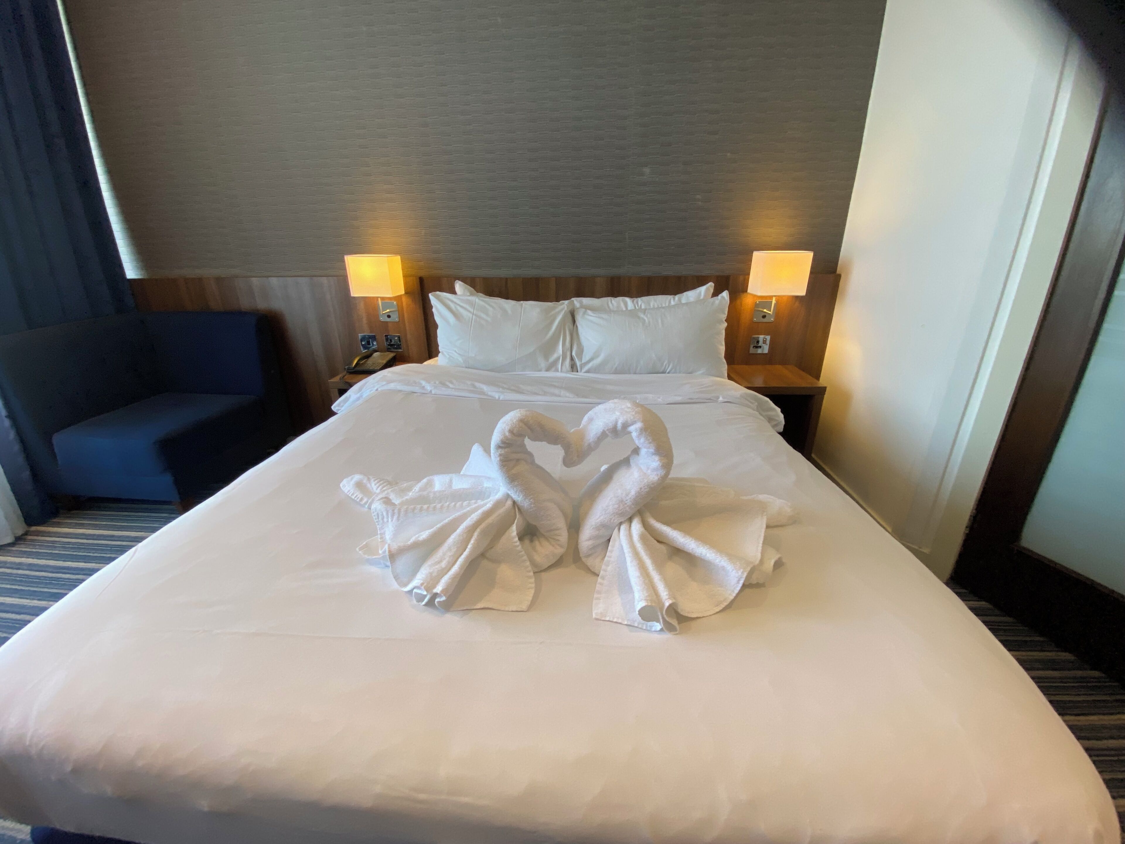 A hotel room with a neatly made bed, two wall-mounted lamps, a blue armchair, and a wooden headboard.