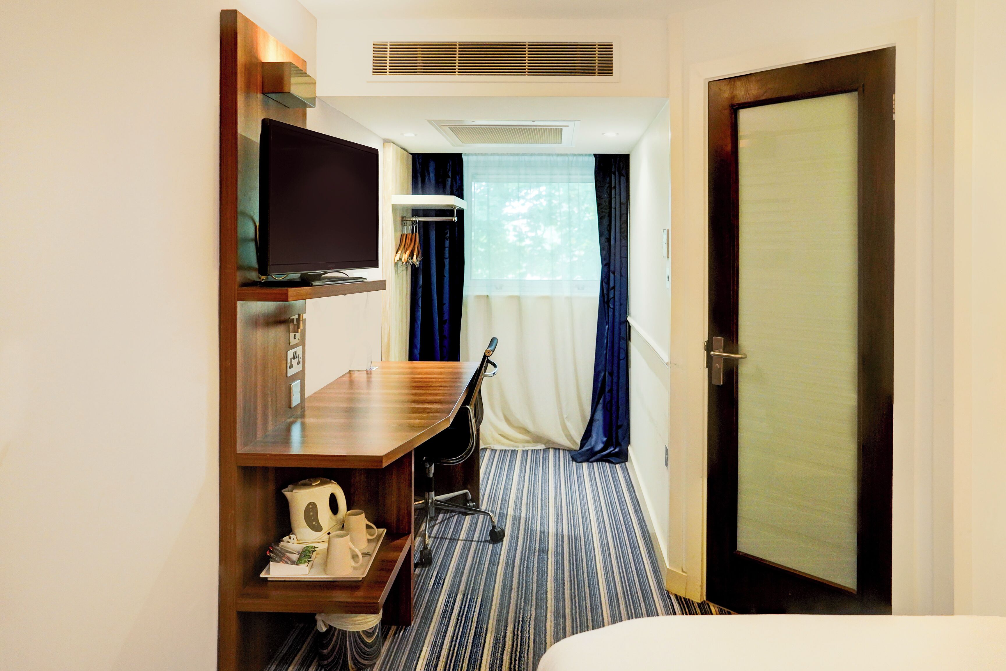 A hotel room with a wooden desk, a flat-screen TV, a bed, and a door.