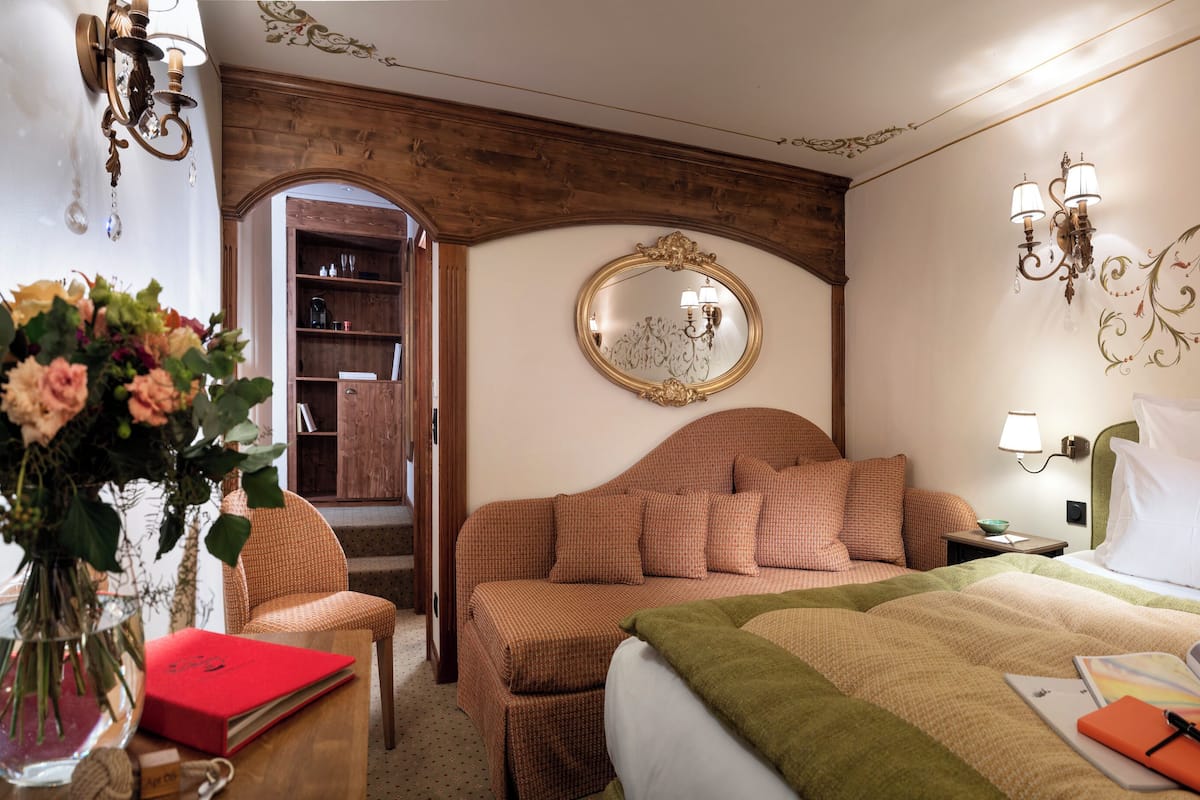 A hotel room with a bed, a bedside table with a lamp, a mirror, a bookshelf, and a vase with flowers.