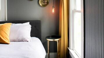 Una cama bien hecha con ropa de cama blanca y gris, una mesita redonda con base dorada y dos platos decorativos en la pared.