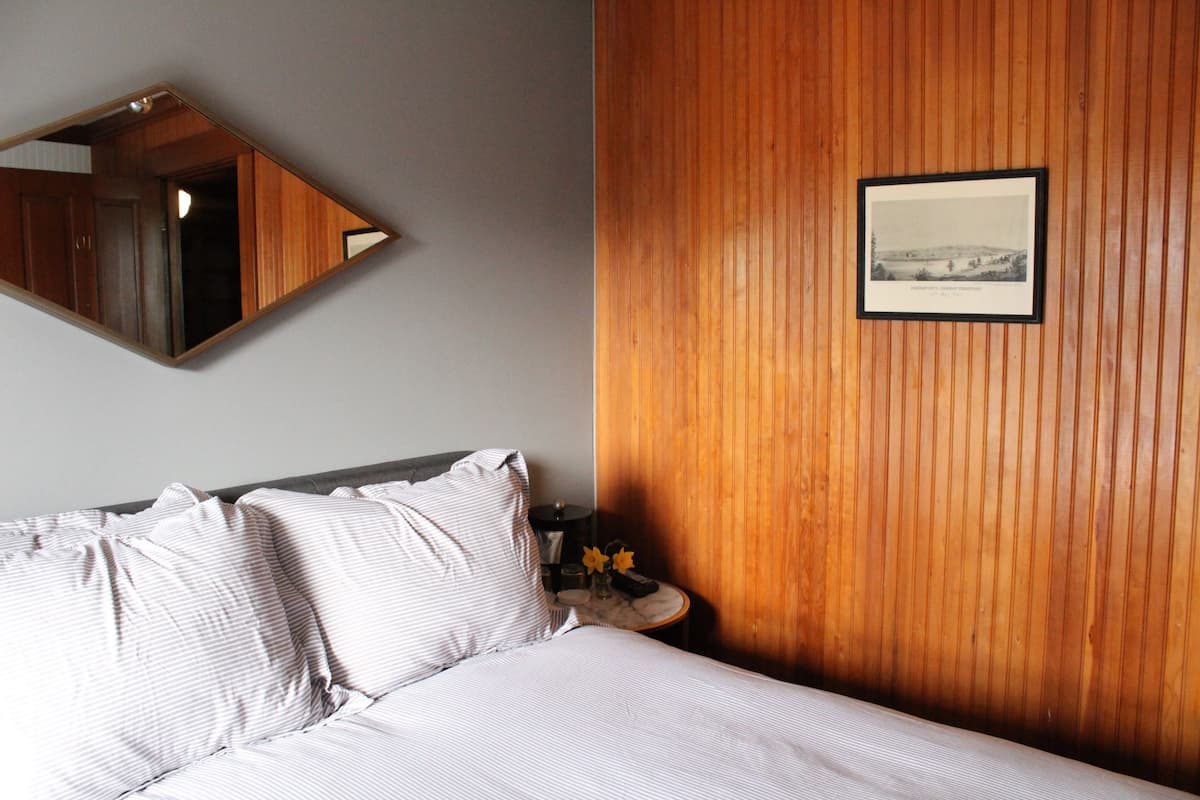 A hotel room with a bed, a wooden panel wall, a framed picture, and a nightstand with a vase of flowers.