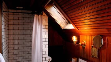 Un baño con techo de madera, cortina de ducha blanca, dispensador de papel higiénico en la pared y un espejo redondo.