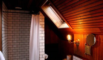 Un baño con techo de madera, cortina de ducha blanca, dispensador de papel higiénico en la pared y un espejo redondo.
