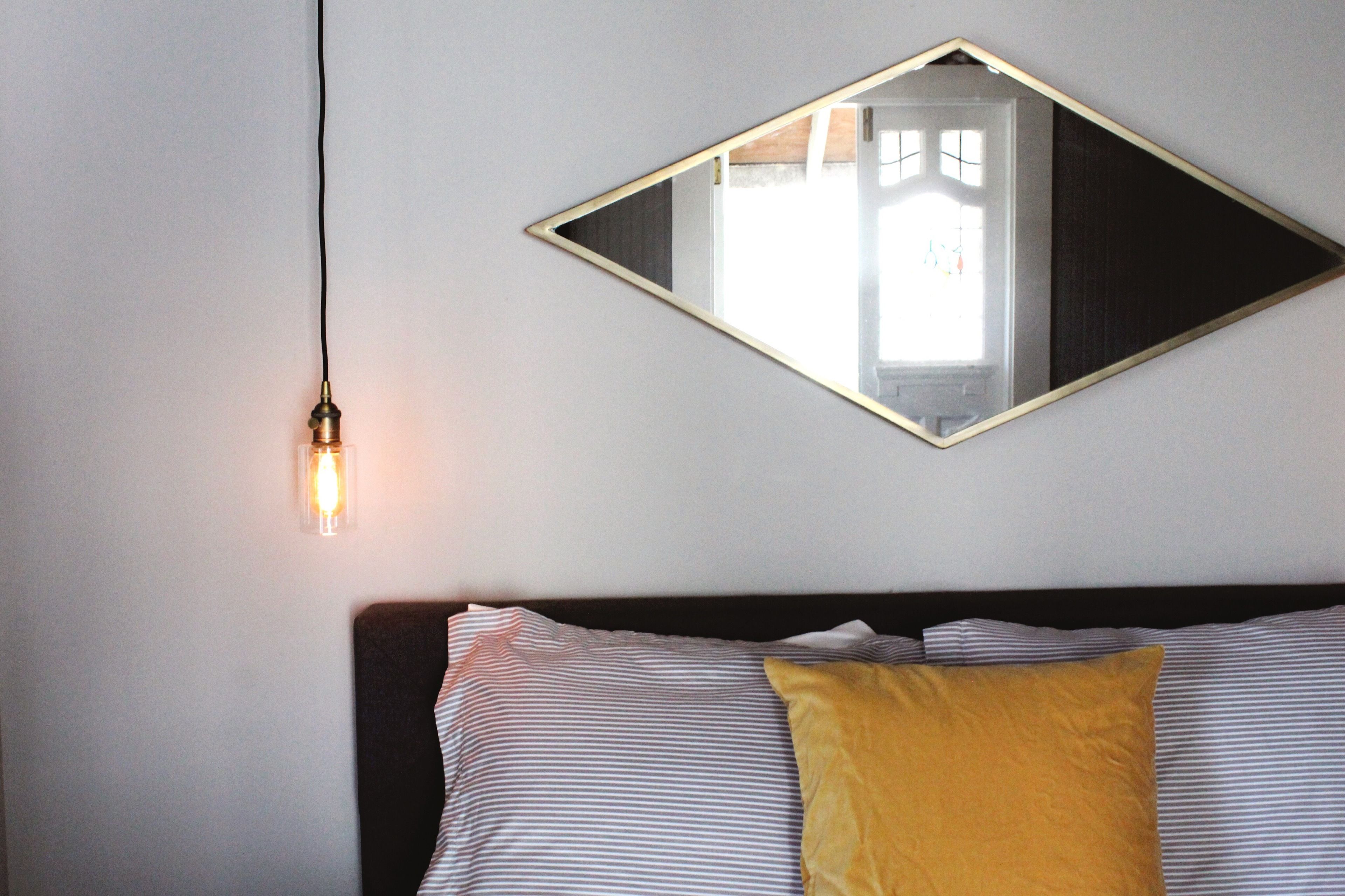 Un lit avec une literie à rayures et un seul oreiller jaune, un miroir fixé au mur et un luminaire suspendu.