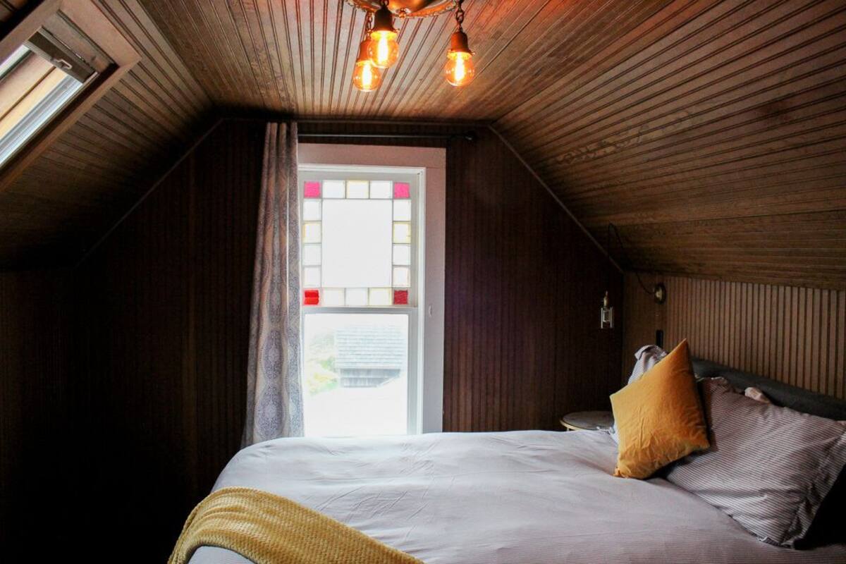 A bedroom with a sloped ceiling, a bed with a white comforter and multiple pillows, a window with a stained glass design, and a ceiling light fixture.