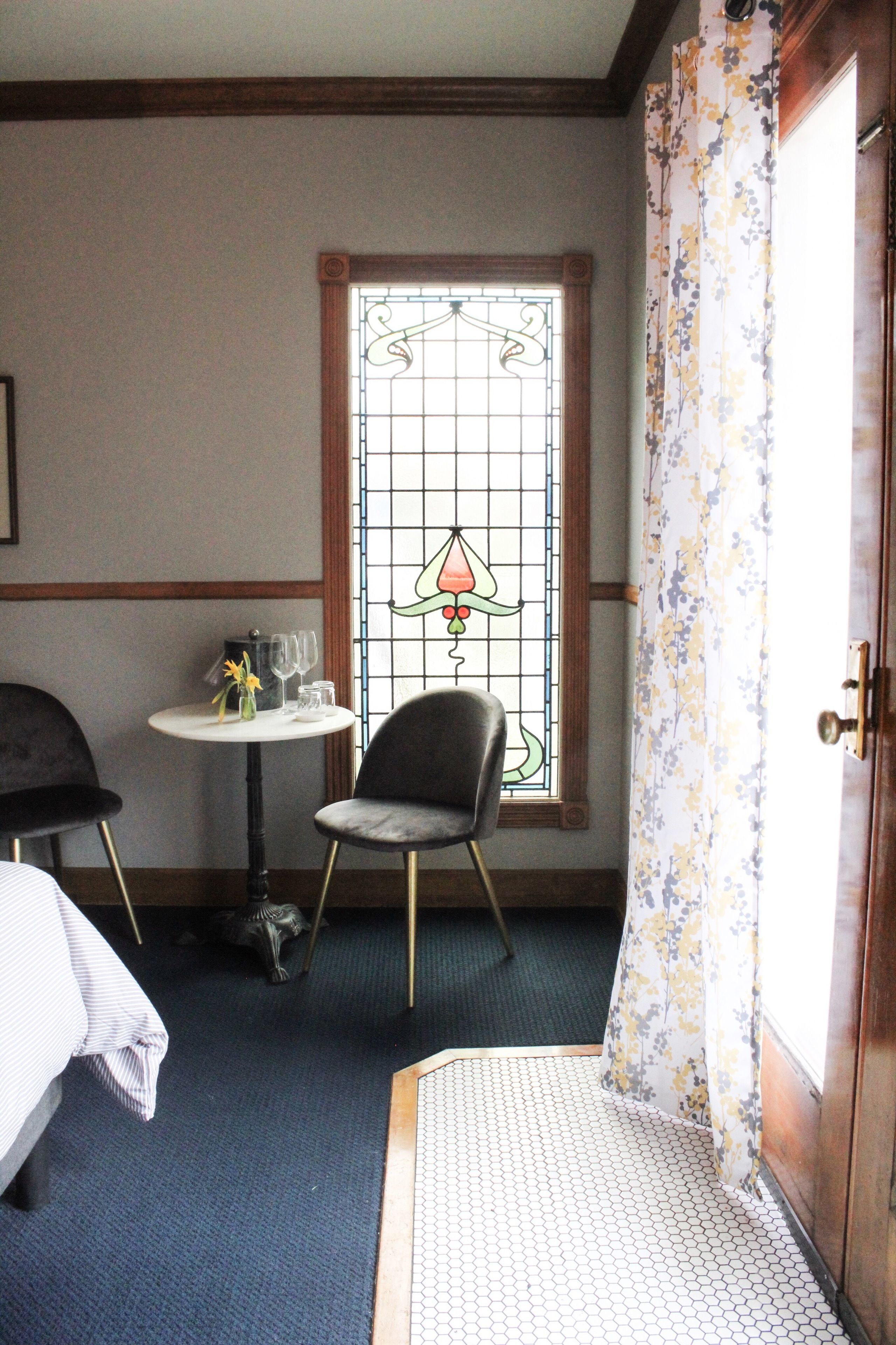 Hotelkamer met een bed, een klein rond tafeltje met een vaas en wijnglazen, een stoel, een raam met een glas-in-lood ontwerp, en een deur met bloemen gordijnen.