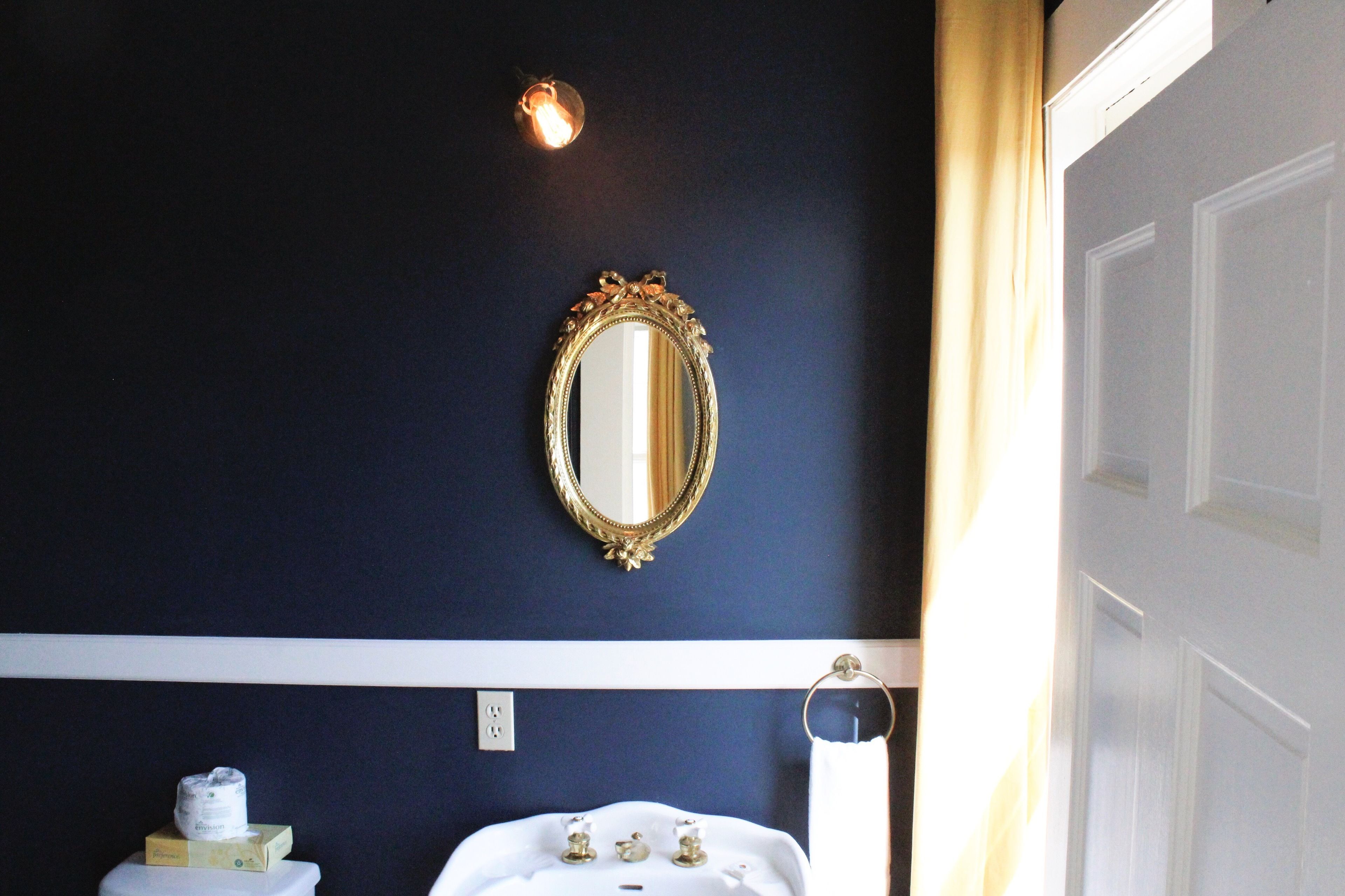A bathroom with a dark blue wall, a gold-framed mirror, a white sink, and a towel rack.