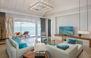 A modern living room with a glass coffee table, two sofas with blue cushions, a wooden side table, a TV on a wooden stand, and a view of the ocean through large windows.