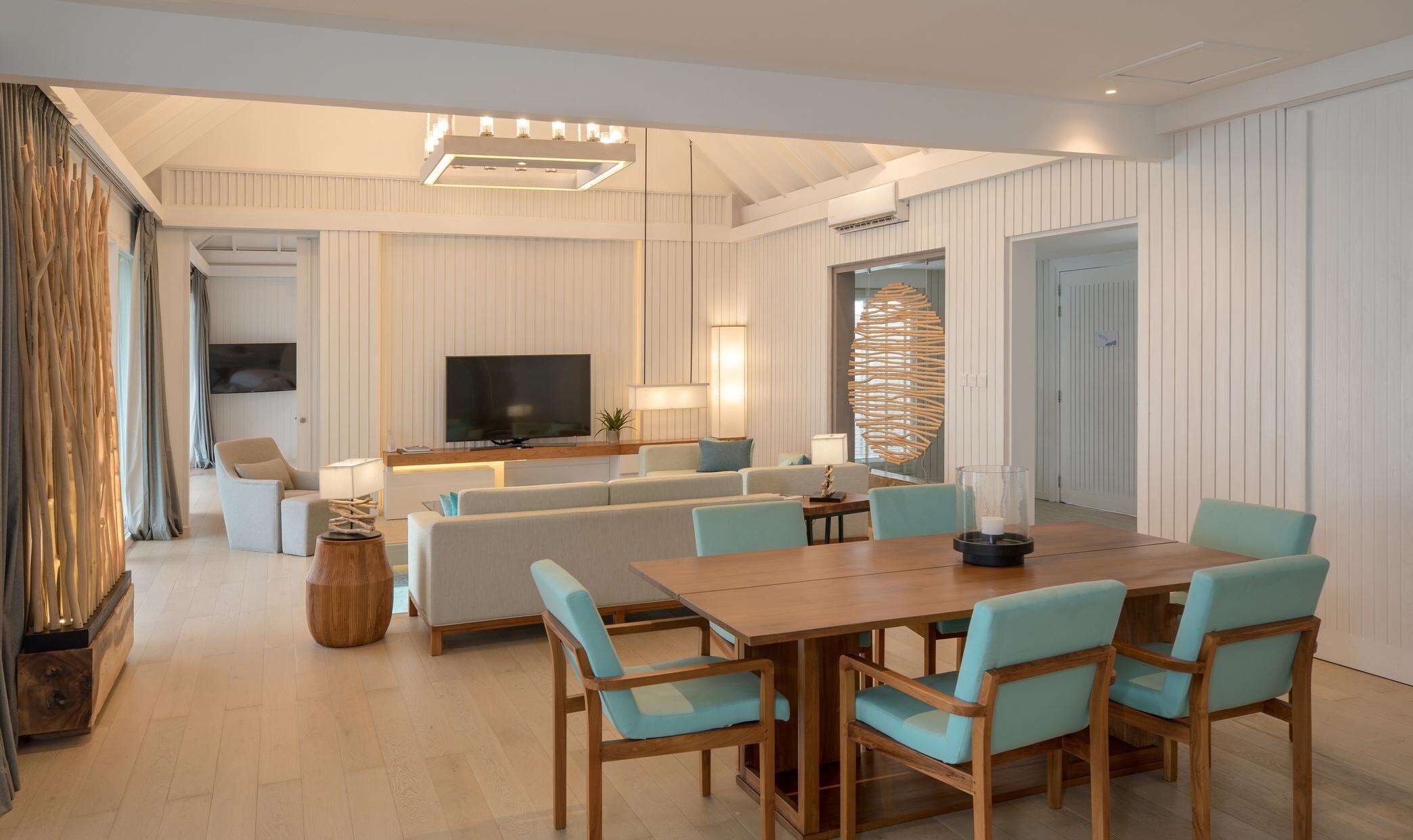 A modern living room with a wooden dining table, light blue upholstered chairs, a sofa, and a television.
