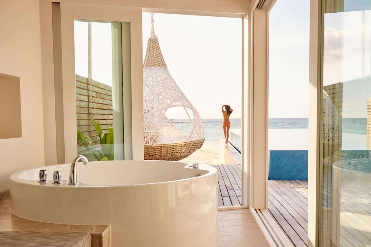 Ein modernes Badezimmer mit einer großen Badewanne, einem Korbstuhl und Blick auf den Strand.