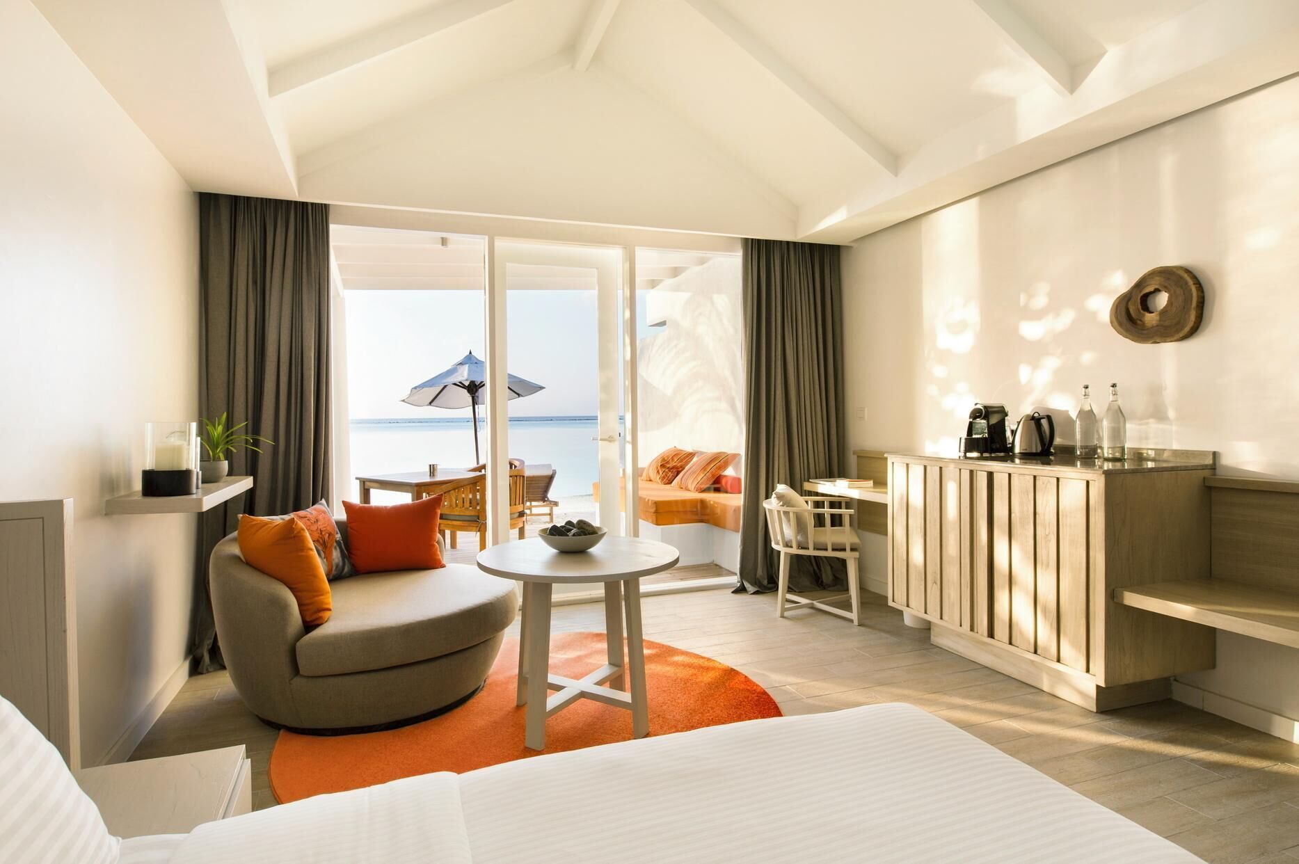 A hotel room with a bed, a seating area with a table, a desk with a chair, a minibar, and a view of the sea.