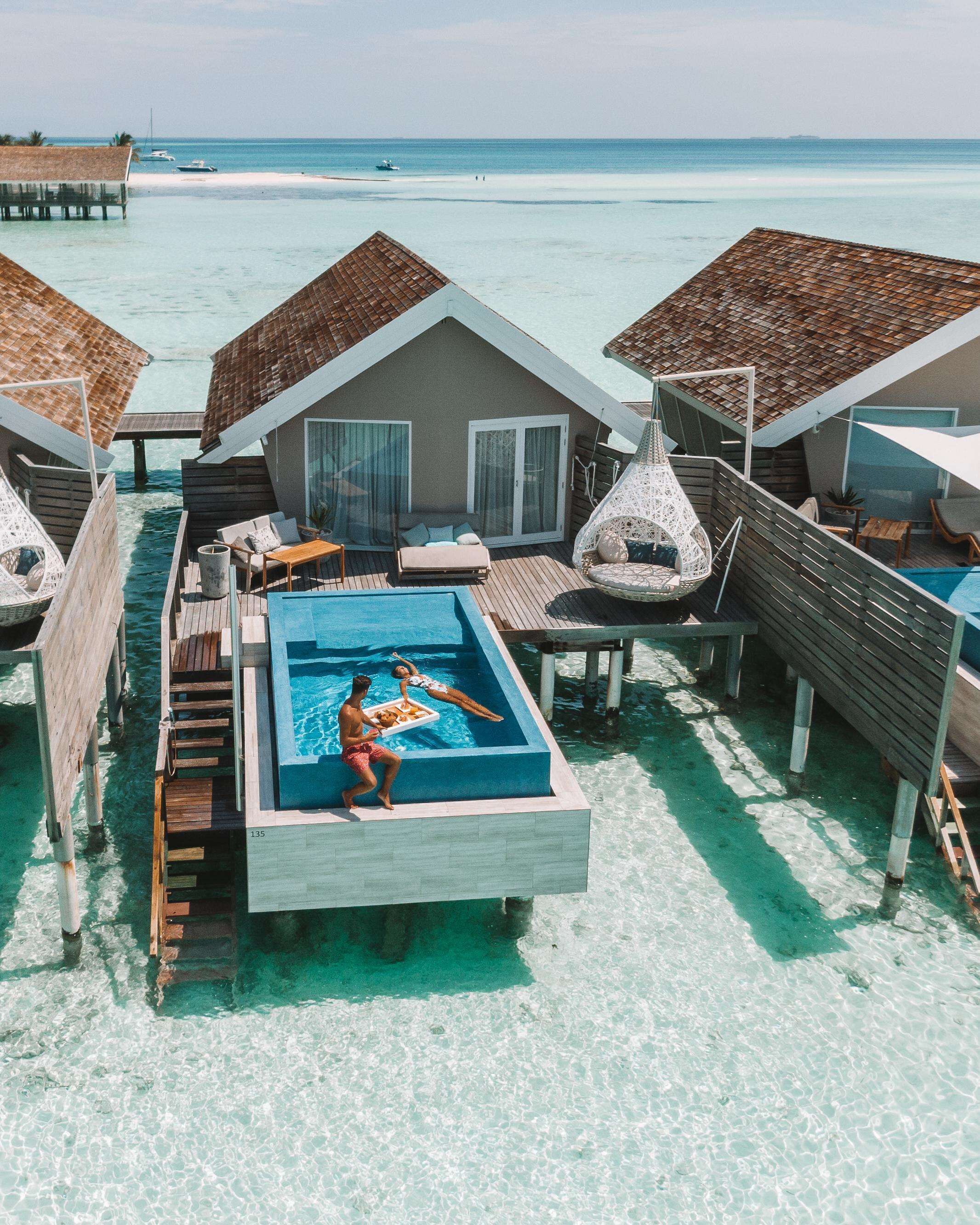 Ein Bungalow auf Stelzen über dem Wasser, mit Pool, Holzterrasse und strohgedeckten Strukturen.