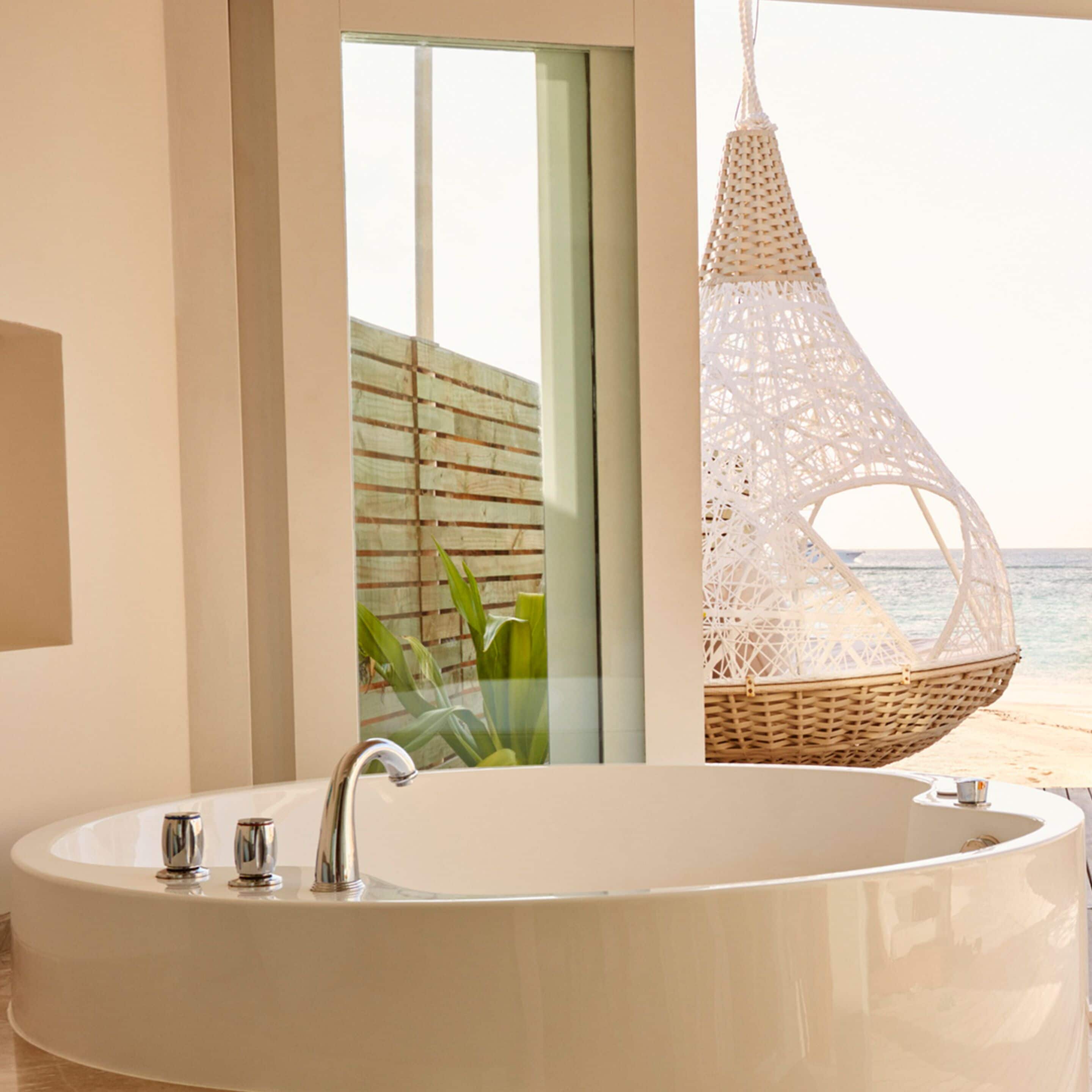 A modern bathroom with a large bathtub, a wicker hanging chair, and a view of the sea.