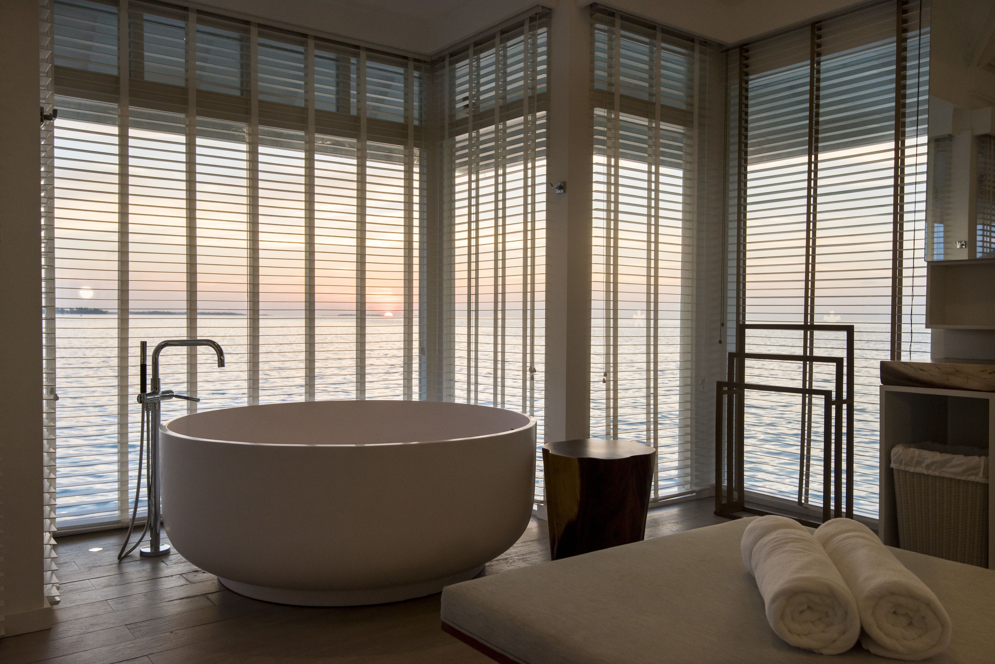 A modern bathroom with a freestanding bathtub, large windows with blinds, and a view of the sea.