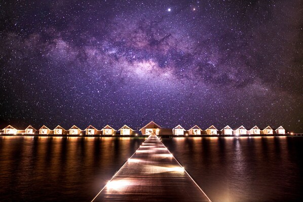 A starry night sky over a wooden pier leading to overwater bungalows.