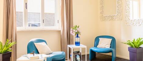 A room with two blue chairs, a small white table with a book, a vase with flowers, and a mirror on the wall.