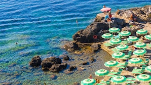 Una playa con agua azul cristalina, costa rocosa y varias sombrillas verdes.