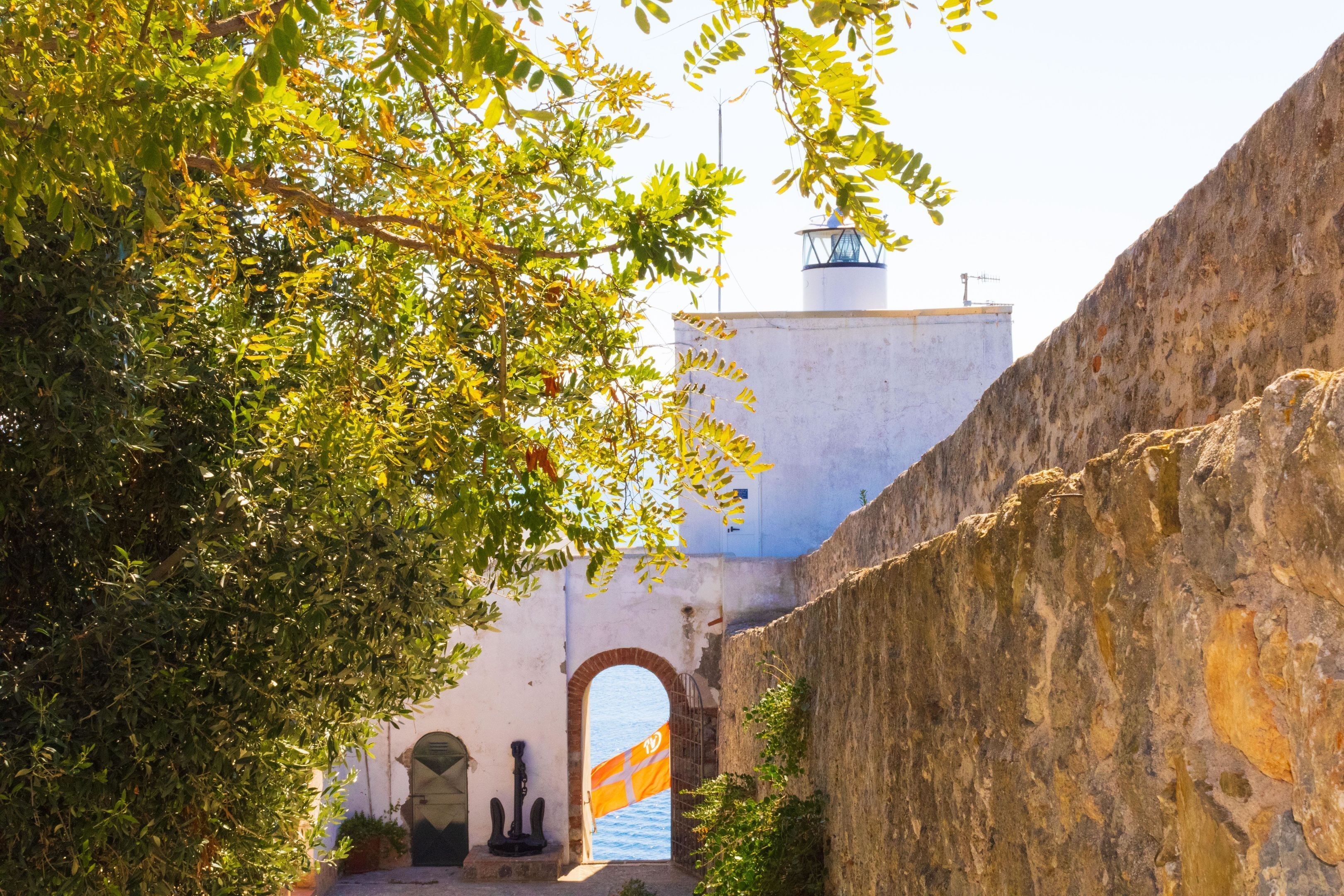 Une ruelle étroite, bordée d’un mur de pierre, avec une porte en arc de cercle et une vue sur la mer par une fenêtre.