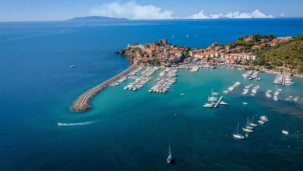 Vista aérea de un pueblo costero con un puerto deportivo lleno de barcos, un faro y edificios históricos.