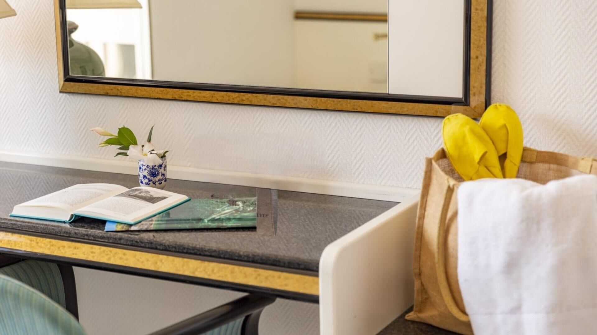 Habitación de hotel con escritorio, espejo, un pequeño jarrón con flores y un racimo de plátanos.