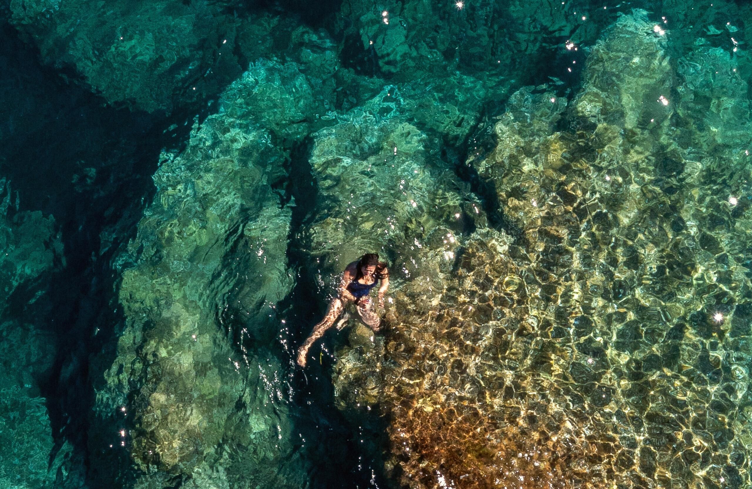 A person swimming in clear, shallow water with rocky formations.