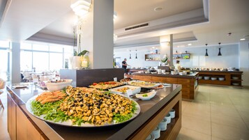 Buffet com diversos pratos, incluindo frutos do mar e saladas, numa área de refeições moderna com amplas janelas e disposição limpa e organizada.