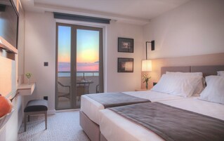 Quarto de hotel com duas camas, uma escrivaninha com cadeira, uma mesinha com um vaso e vista para o pôr do sol pelas portas da varanda.