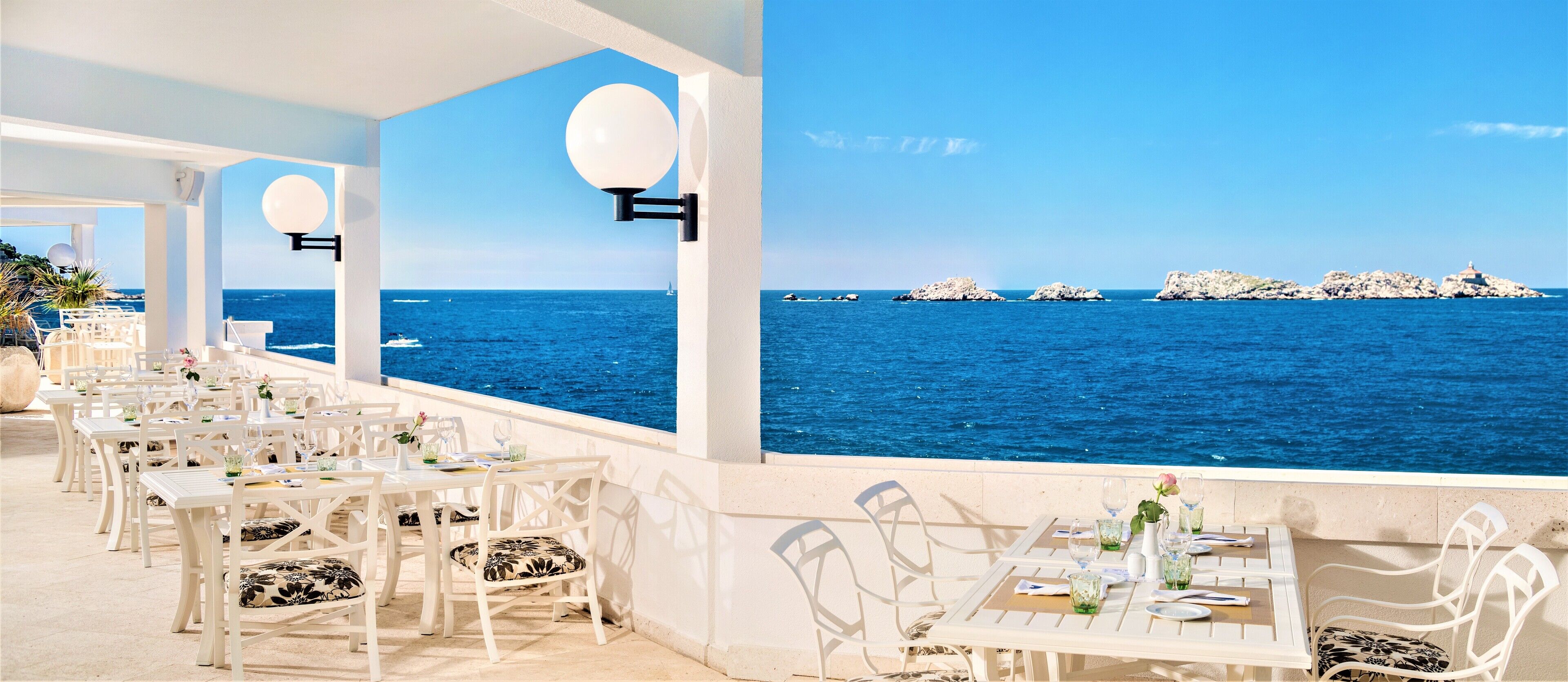A seaside restaurant with white tables and chairs, a clear view of the ocean, and rocky formations in the distance.