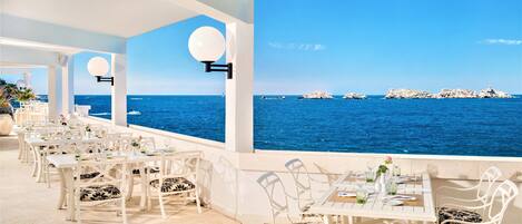 A seaside restaurant with white tables and chairs, a clear view of the ocean, and rocky formations in the distance.