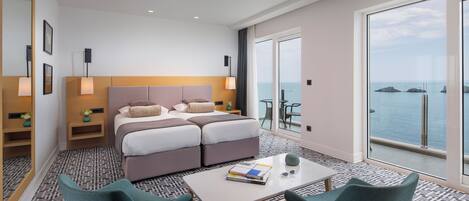 Ein modernes Hotelzimmer mit einem großen Bett, zwei Nachttischen, einem kleinen Tisch mit Büchern, zwei Stühlen und einem großen Fenster mit Meerblick.