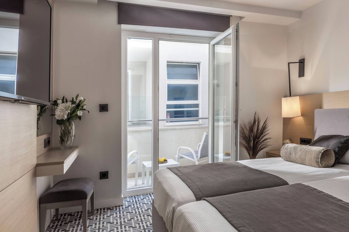 Une chambre d’hôtel moderne avec un balcon, un lit avec une literie grise, une table de chevet et un téléviseur fixé au mur.