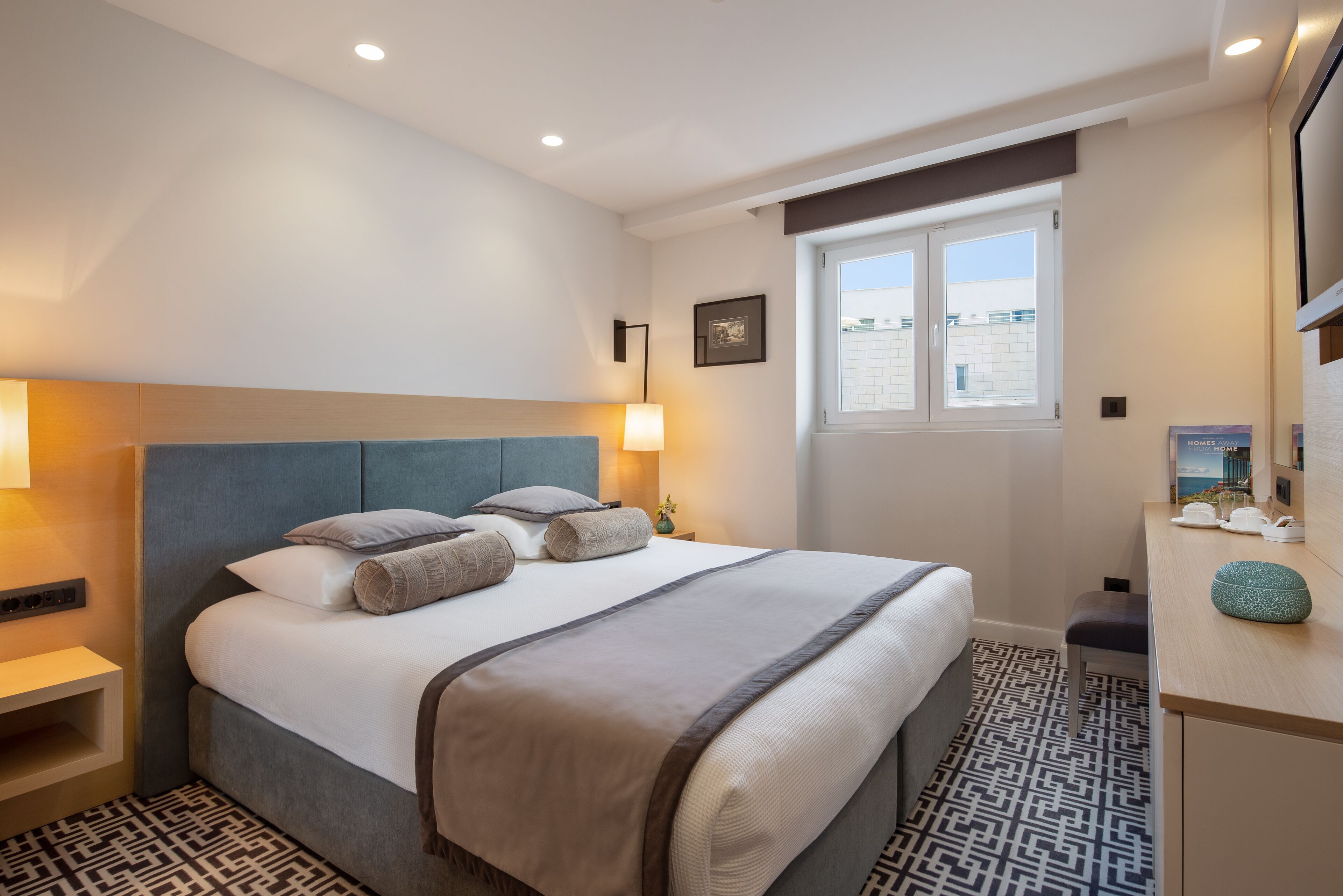 Habitación de hotel con una cama grande, mesitas de noche, un escritorio con silla, una ventana y vistas a edificios.