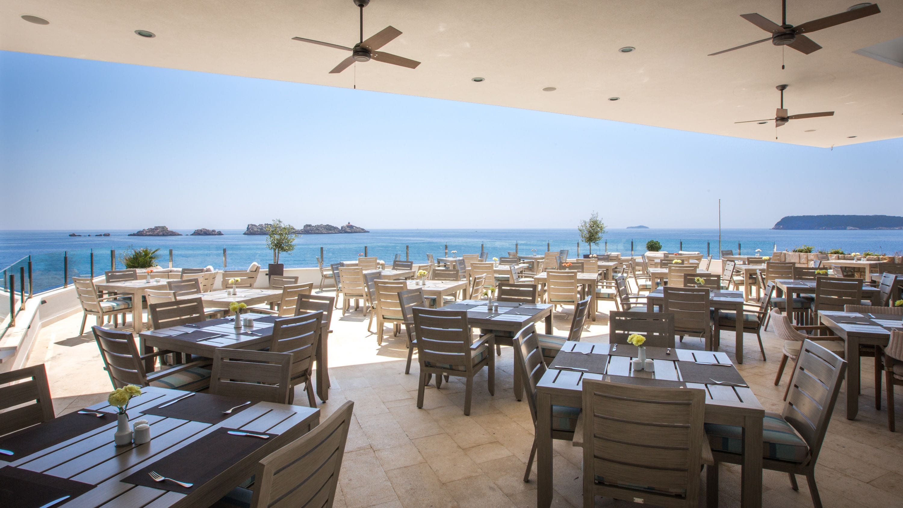 A seaside restaurant with multiple tables and chairs, ceiling fans, and a view of the ocean.