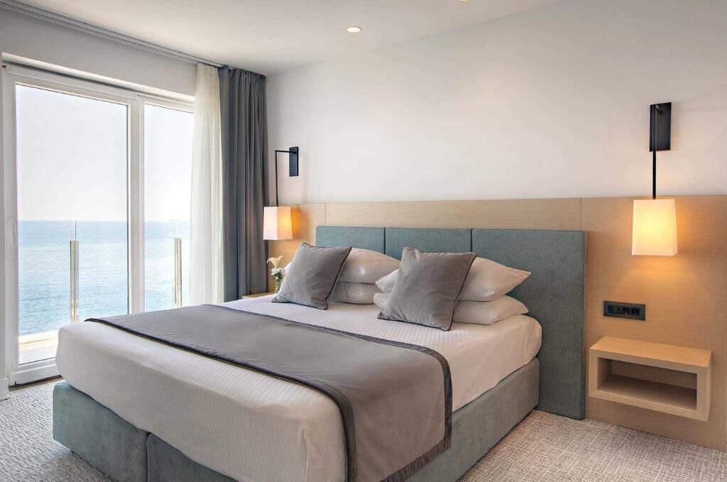 Une chambre d’hôtel avec un grand lit, une table de chevet, une lampe fixée au mur et une vue sur la mer.