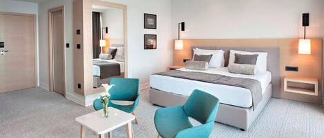 A hotel room with a large bed, two bedside tables, a small table with a vase, two teal chairs, and a mirror.