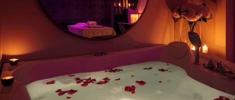 A spa room with a large bathtub filled with water and rose petals, a mirror on the wall, candles, and a vase with flowers.