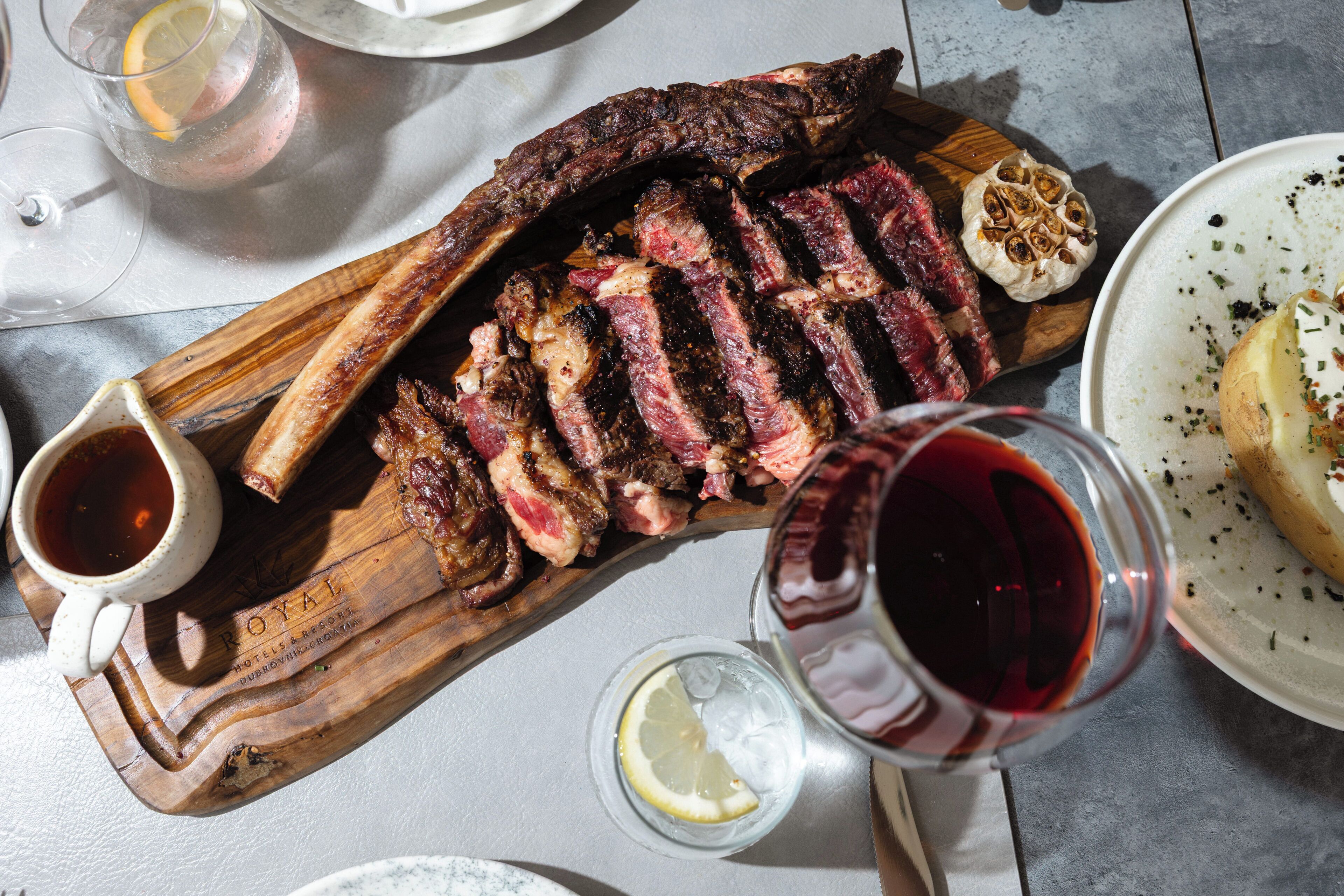 Uma tábua de madeira com carne fatiada, um copo de vinho tinto e uma xícara de molho.