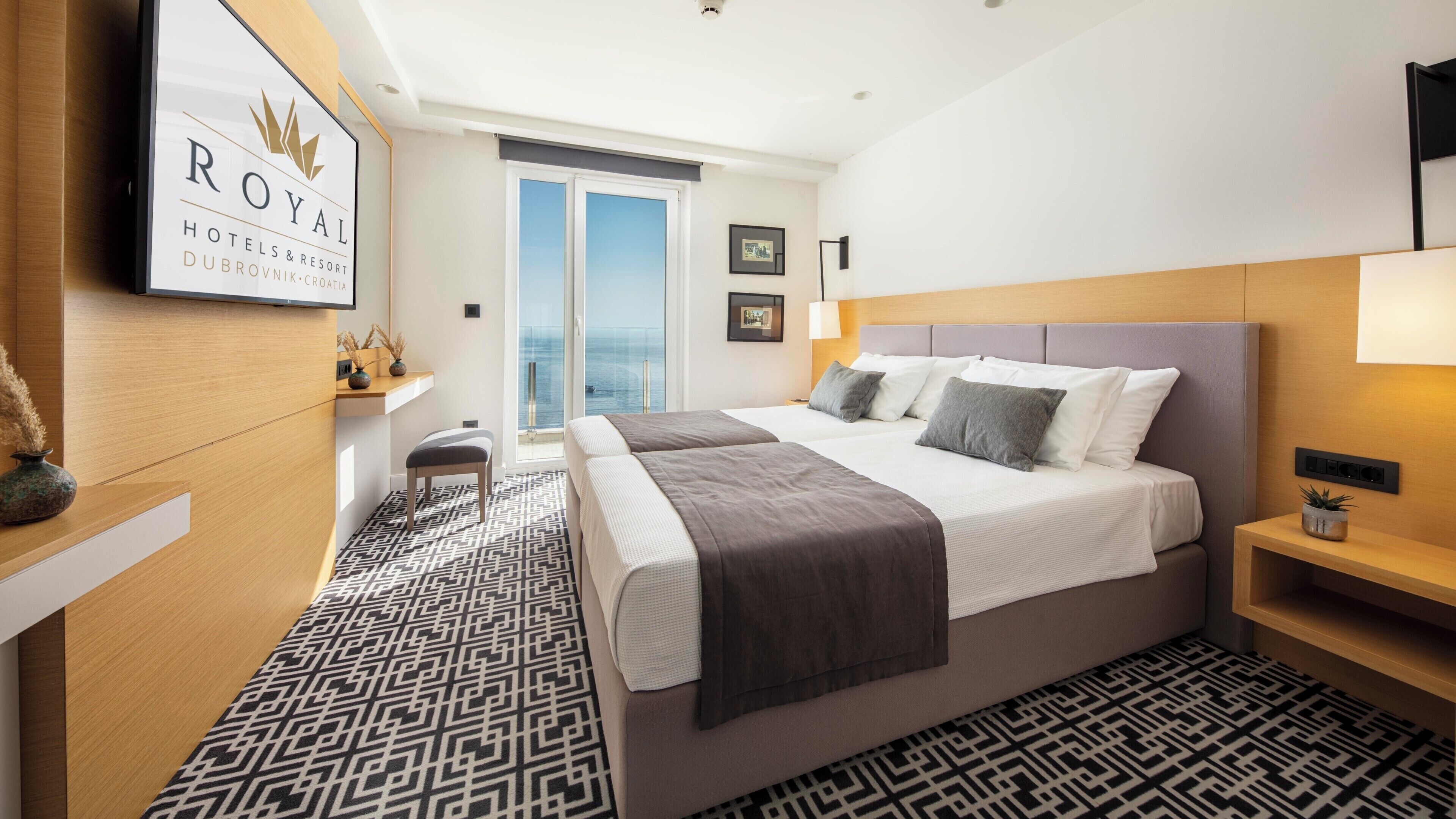 A hotel room with a large bed, a bench, a desk, and a view of the sea.
