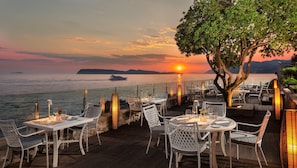 An outdoor dining area with tables and chairs, overlooking a body of water at sunset.