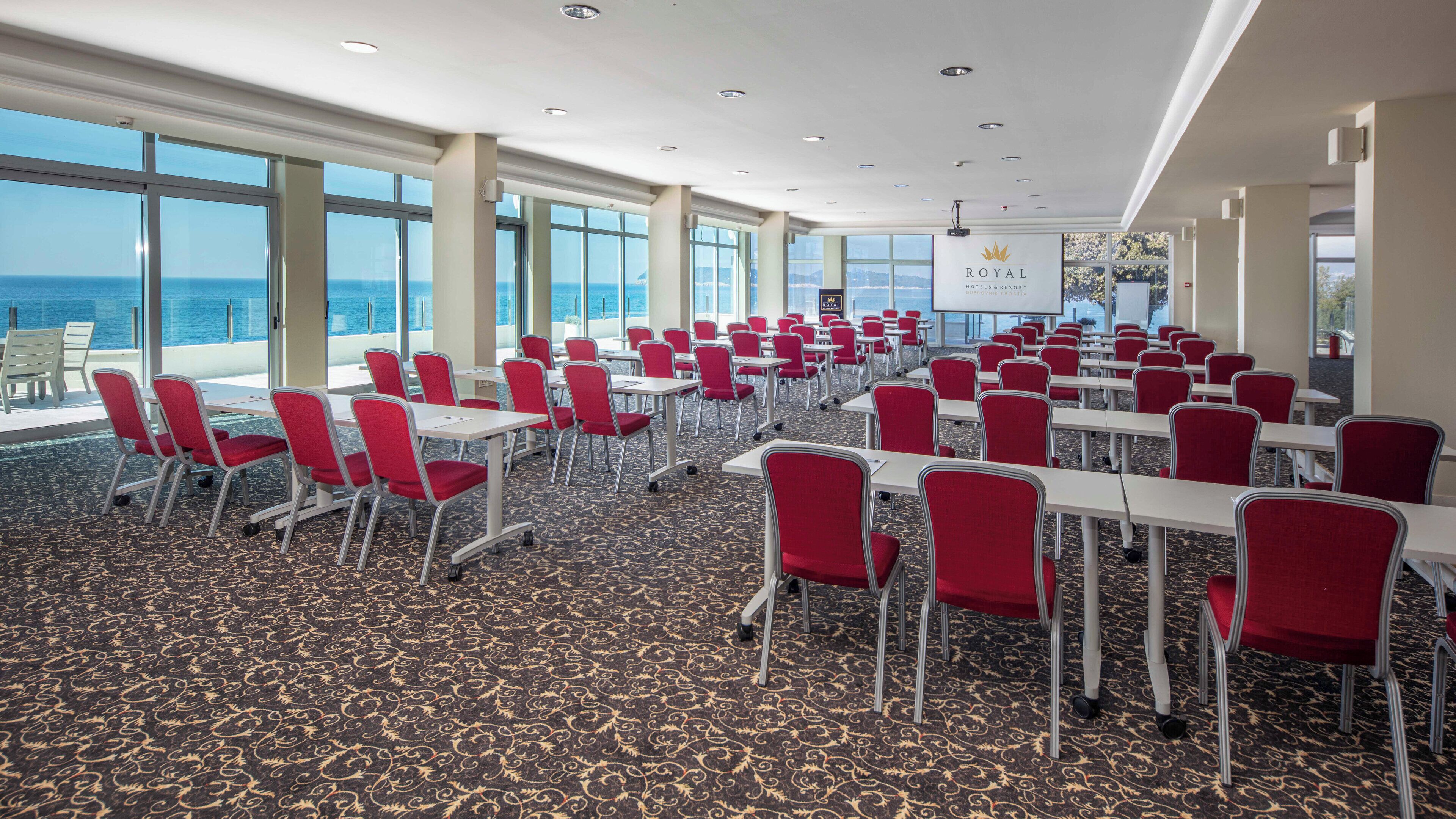 Ein Konferenzraum mit roten Stühlen und Tischen, großen Fenstern mit Meerblick und einem Schild für Royal Hotels & Resort.