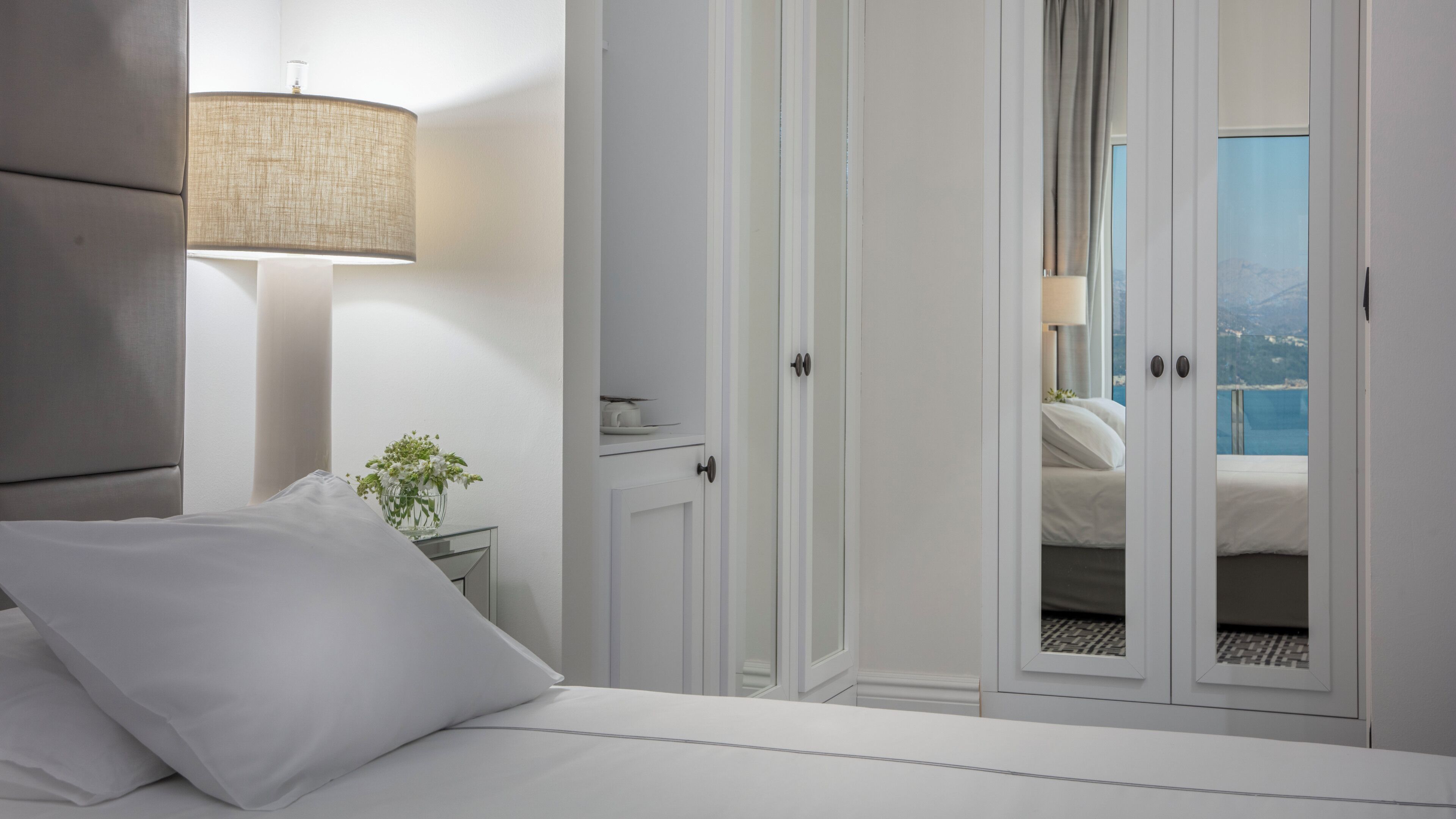 A bedroom with a bed, a nightstand, a lamp, and a view of a balcony with a pool.