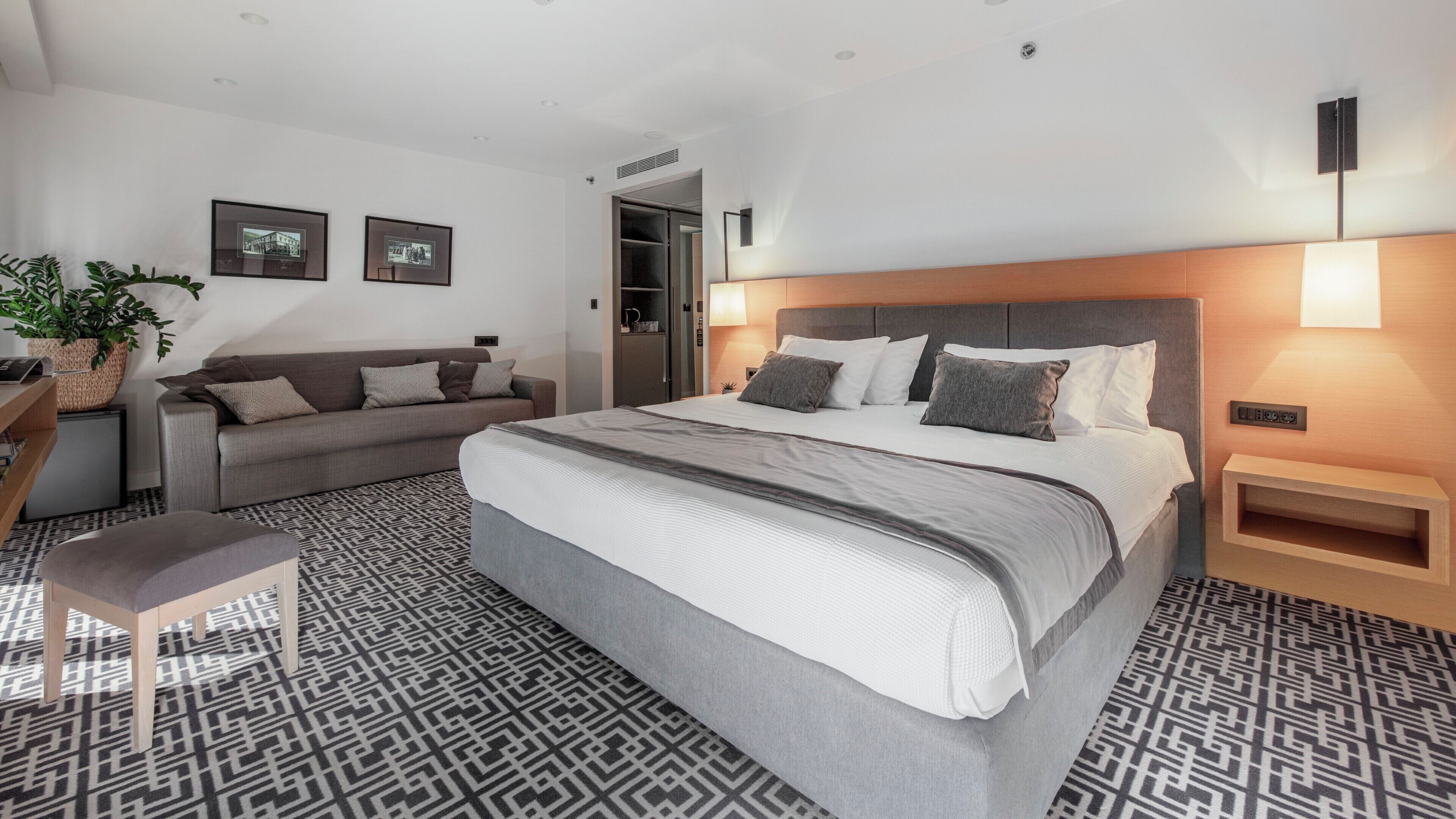 A modern hotel room with a large bed, a sofa, a small table, and a potted plant.