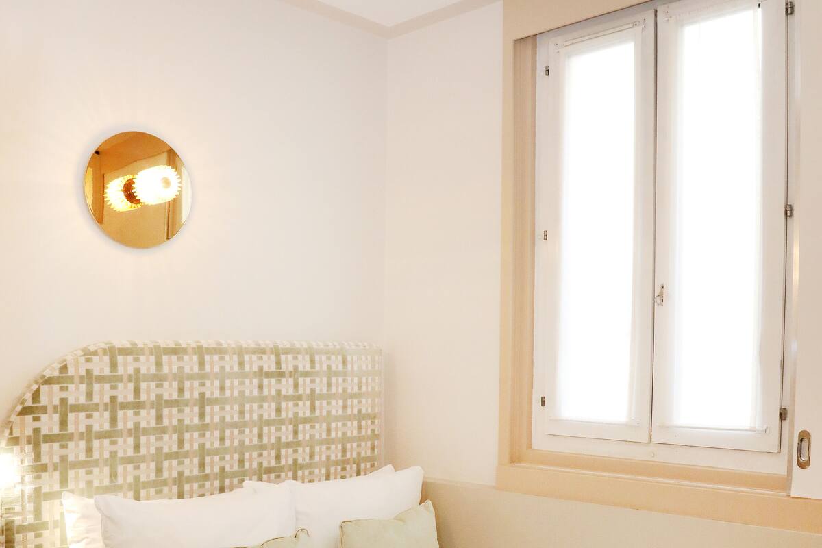 A neatly made bed with a patterned headboard, a bedside lamp, and a window with blinds.