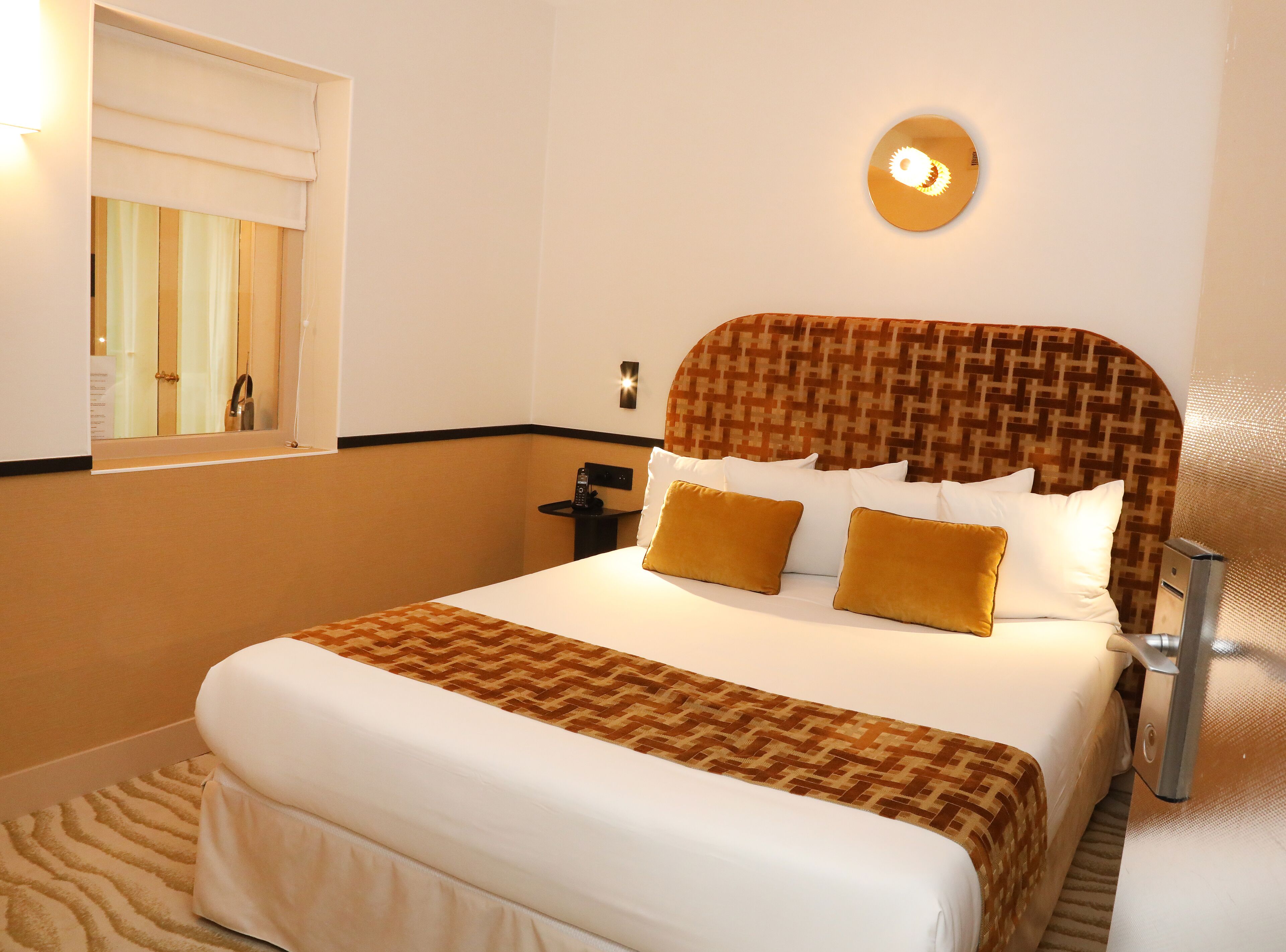 A hotel room with a large bed, a bedside table, a wall-mounted phone, and a window with a view of the outside.