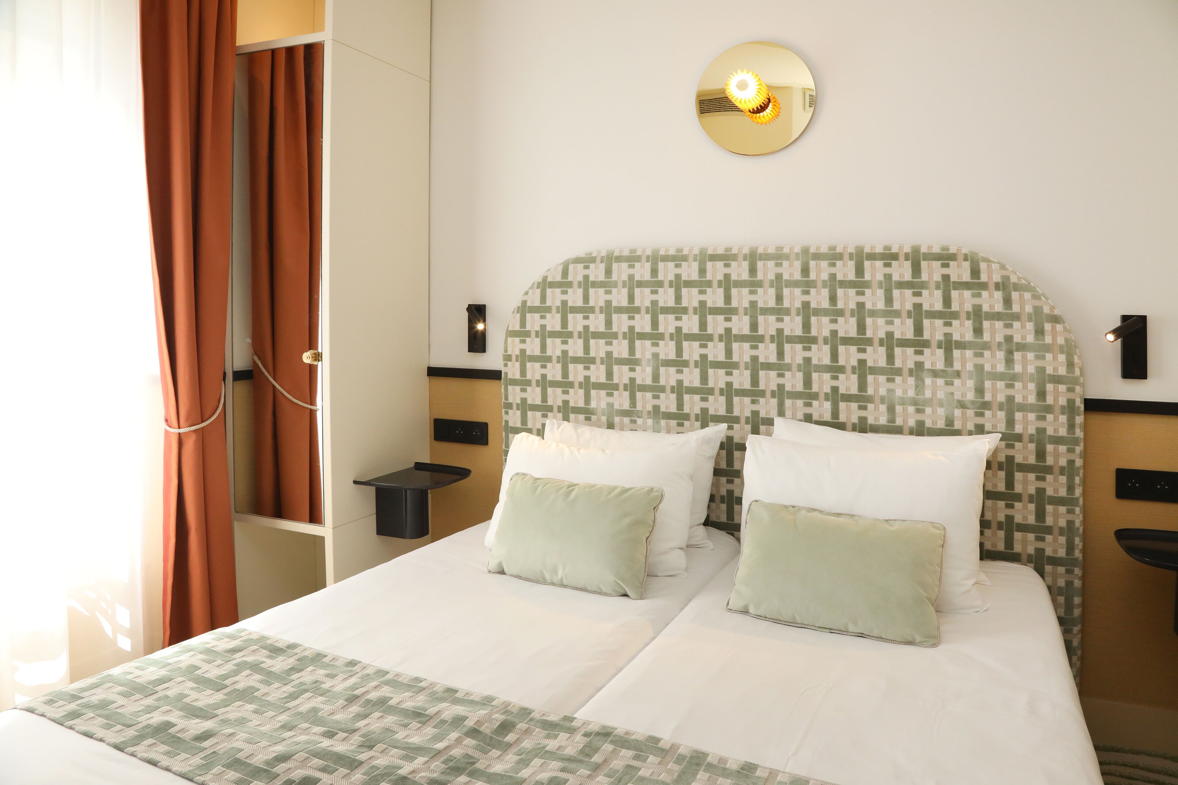 A hotel room with a bed, two bedside tables, a wall-mounted light fixture, and a window with curtains.