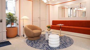 En modern hotellobby med en beige fåtölj, en vit pall, en rund matta, ett litet bord med en vas och en röd soffa.