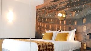 Ett modernt hotellrum med en säng, en väggmålning som föreställer en stadsbild, en hängande lampa och en mönstrad matta.
