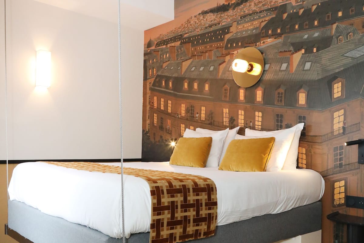 Una habitación de hotel moderna con una cama, un mural de paisaje urbano, una lámpara colgante y una alfombra estampada.