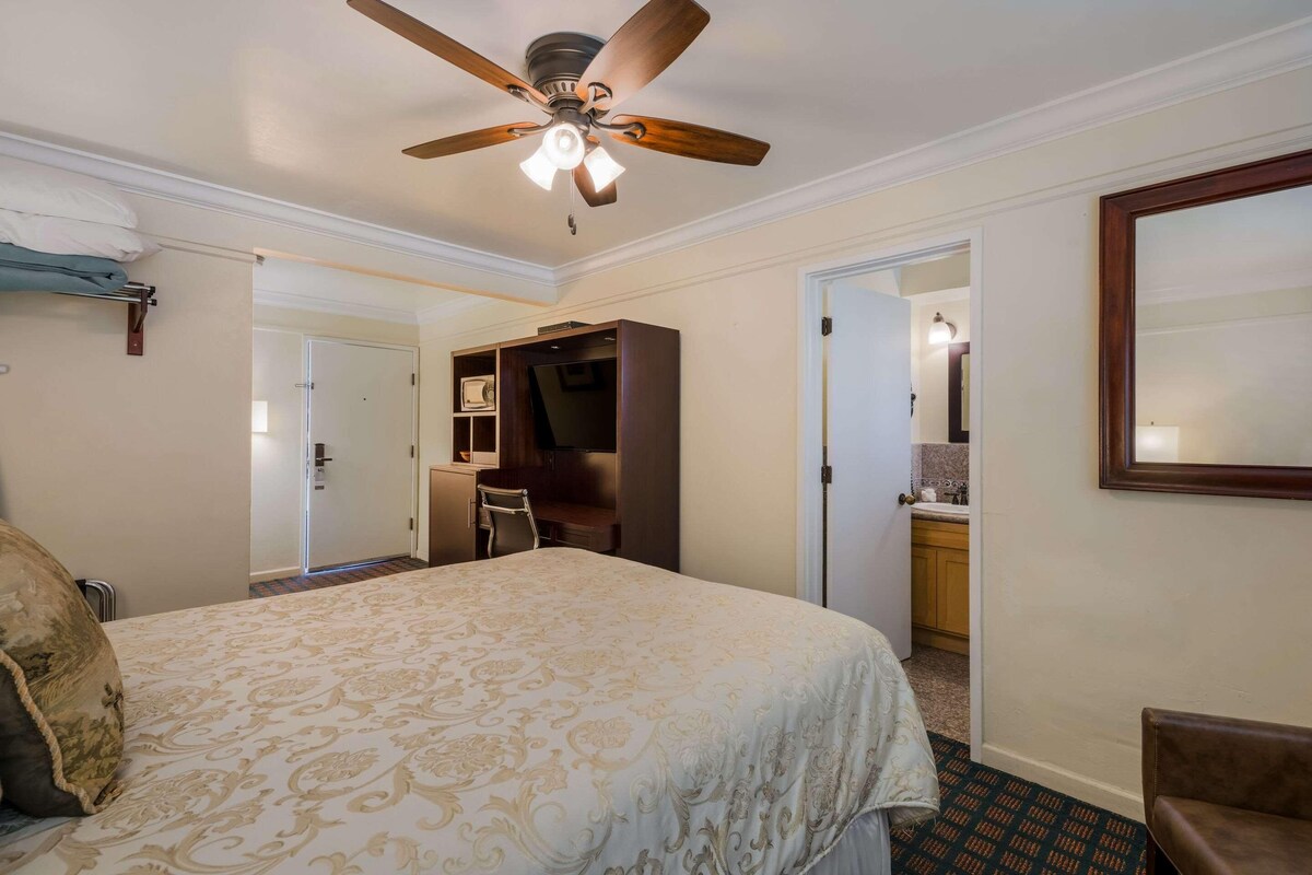 Una habitación de hotel con cama, escritorio, silla, armario y ventilador de techo.